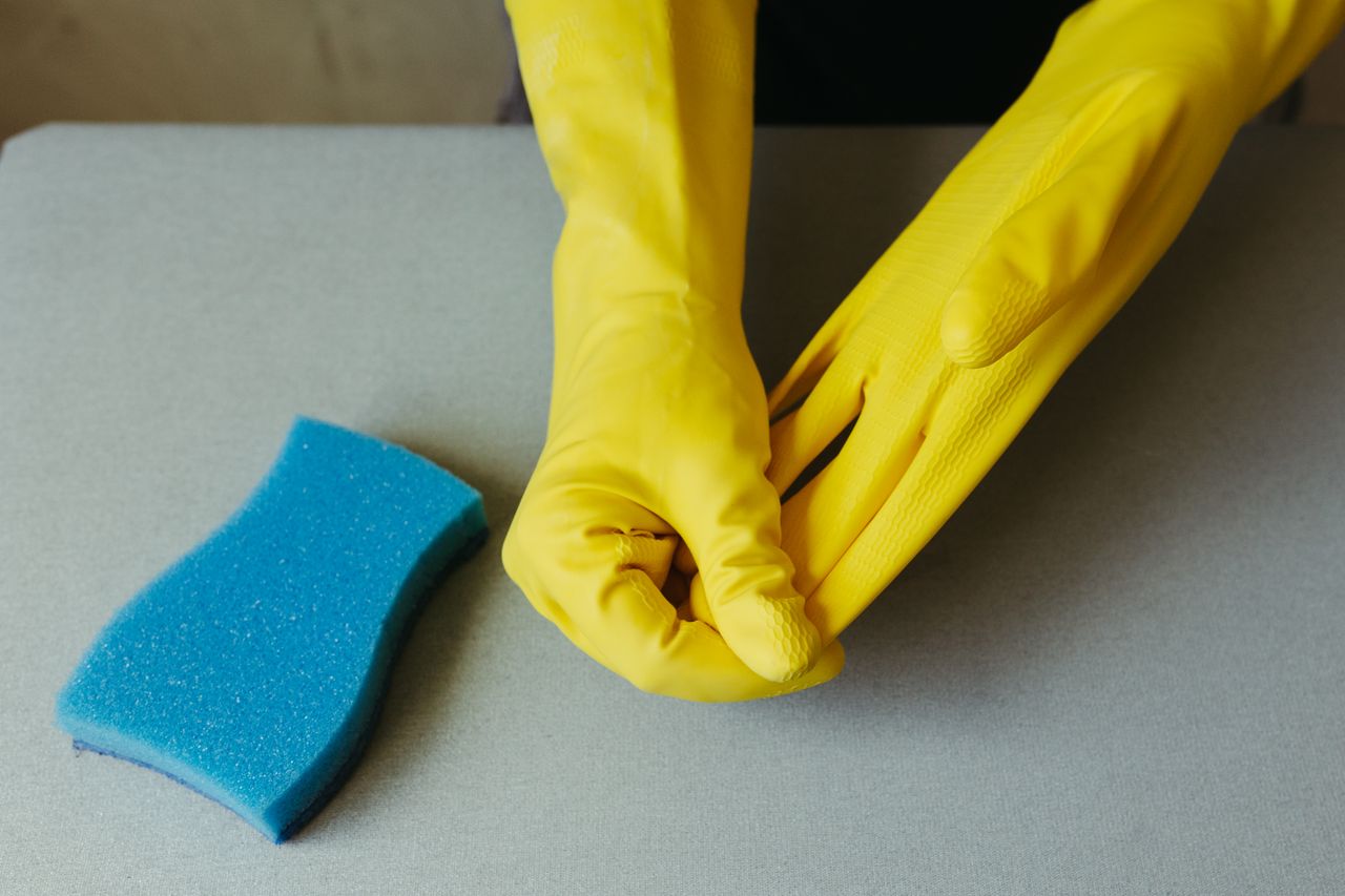 hero-img-01 Close-up of hands wearing yellow gloves next to blue sponge on gray surface.